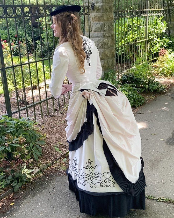19th Century Fashion for Women: 8 Styles and Silhouettes 1 Woman in a cream Victorian dress with a large bustle standing by a fence