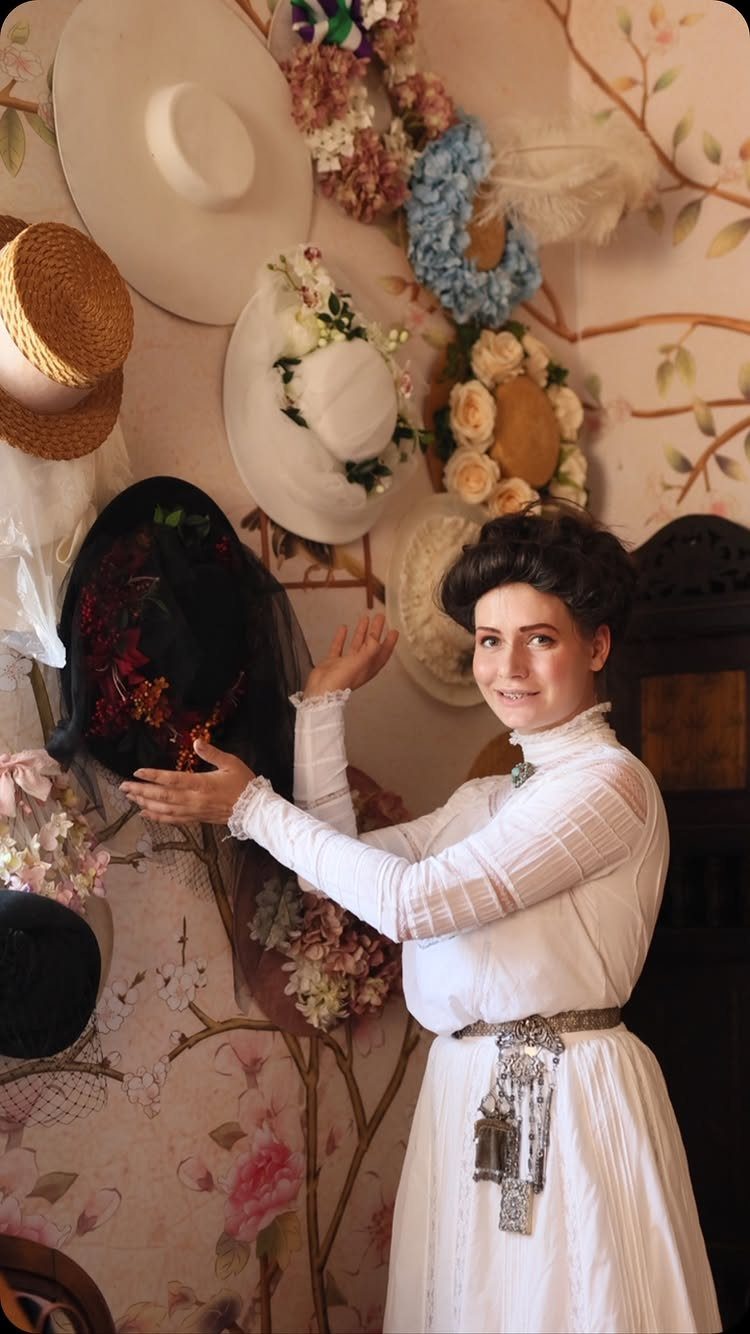 19th Century Fashion for Women: 8 Styles and Silhouettes 8 Woman in white 19th century dress posing by a wall of vintage hats