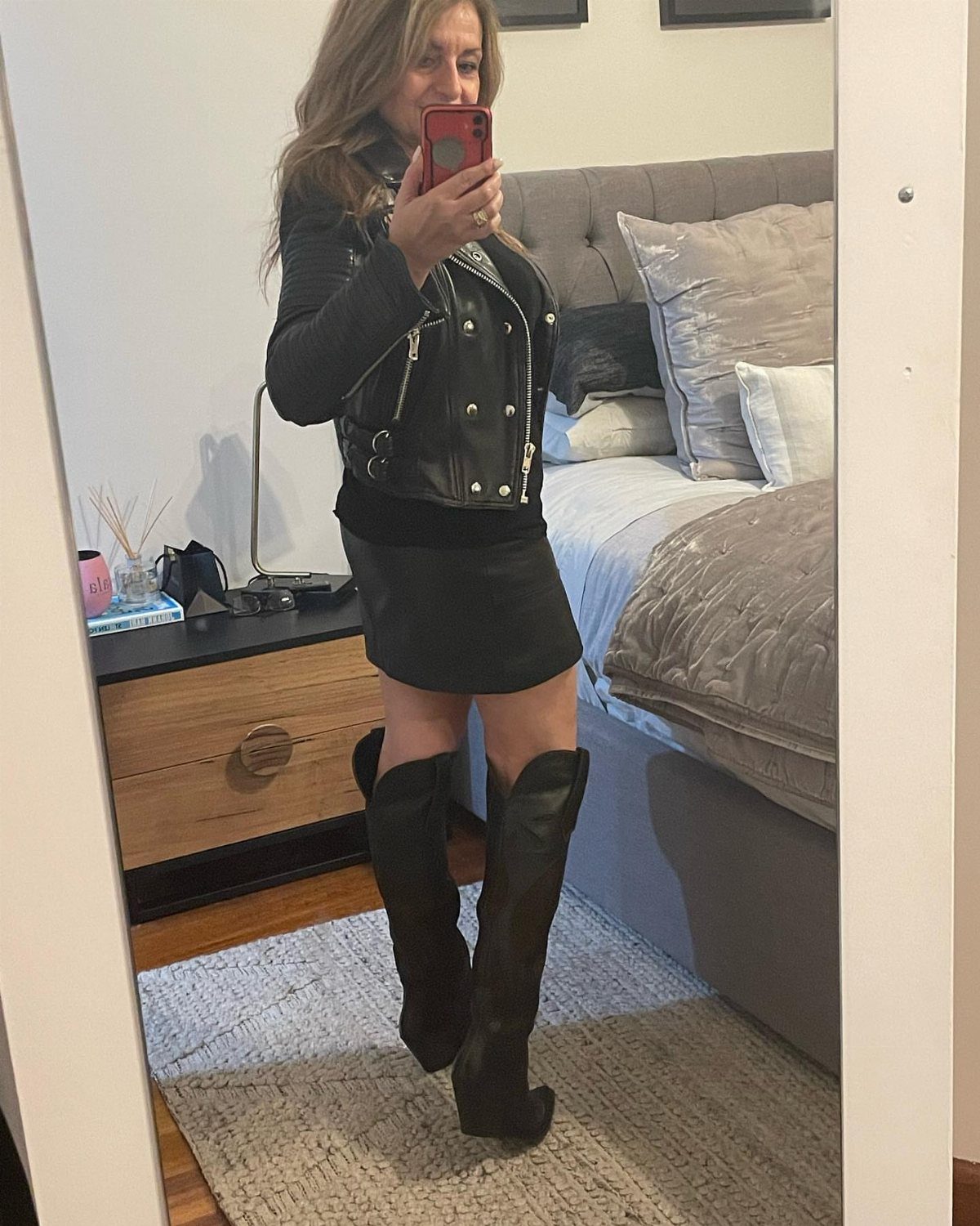 Woman wearing black knee-high boots, a mini skirt, and leather jacket in bedroom.