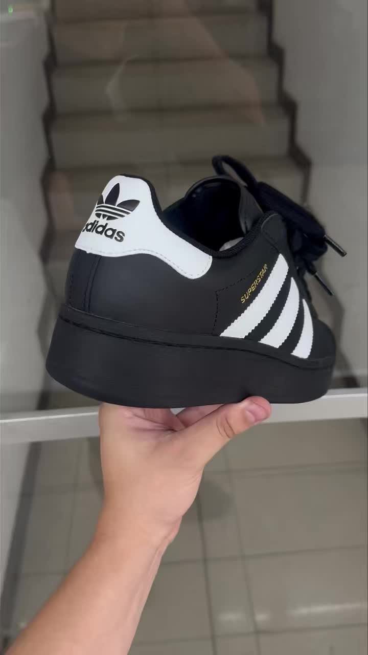 Adidas Superstar Up Wedge with 5 Outfit Ideas to Copy 4 Black Adidas Superstar platform wedge sneaker held by a hand against blurred stairs.