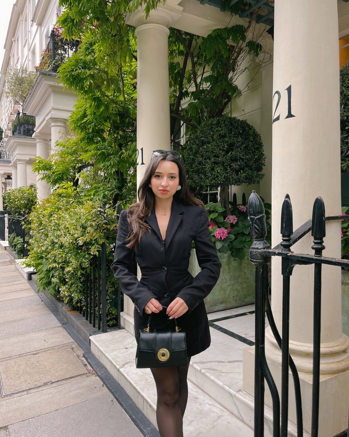 Woman wearing a black blazer dress and tights holding a black handbag outdoors.