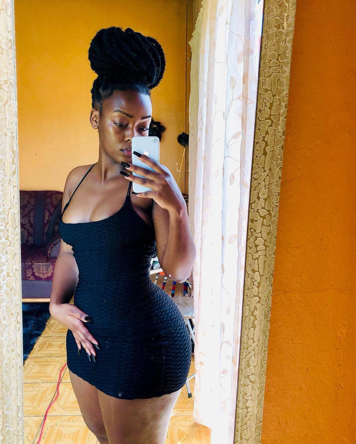 Woman in a tight black mini dress taking a mirror selfie in a room.