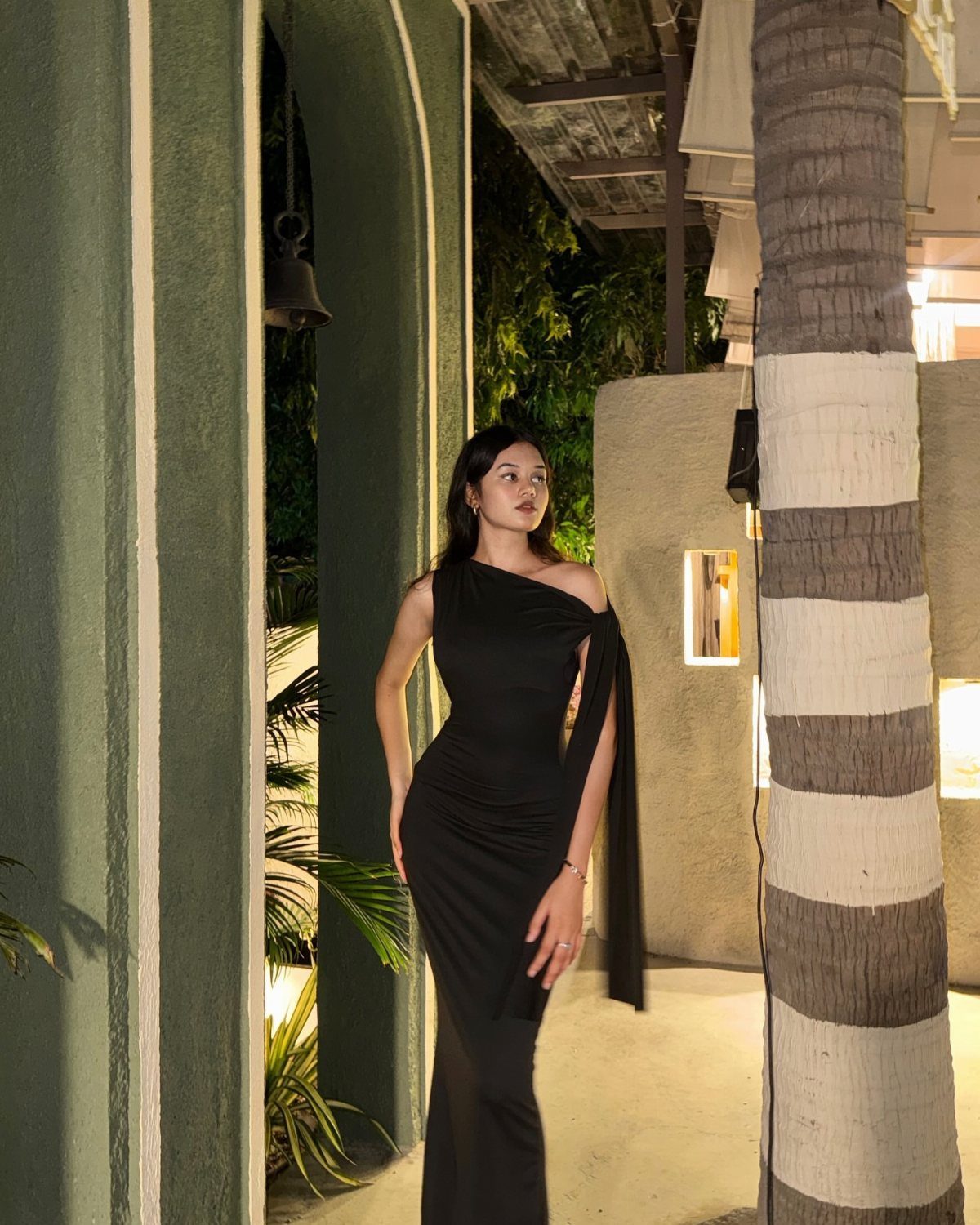 Woman in a one-shoulder black bodycon maxi dress standing outdoors at night.