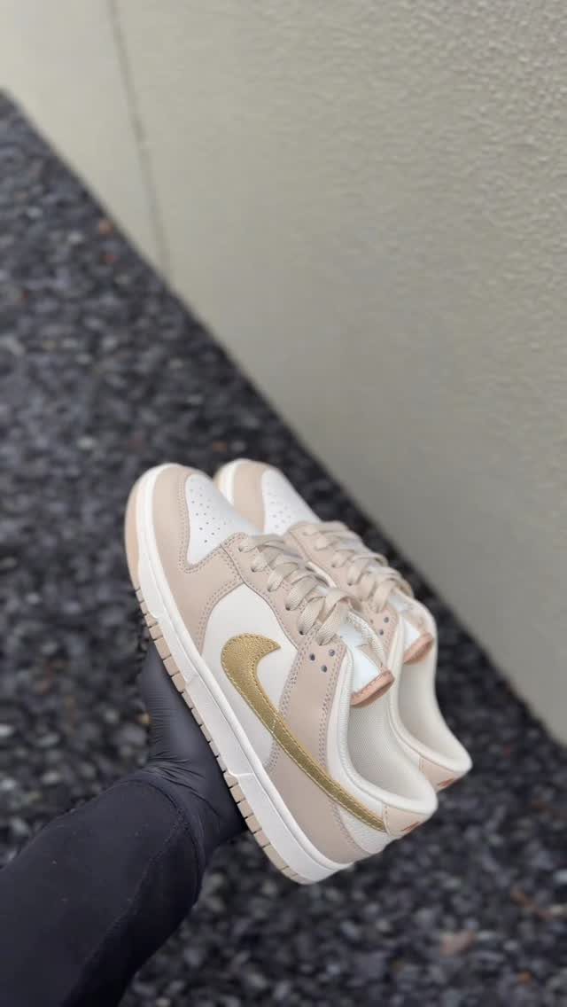 Beige Nike Dunk Low sneakers with metallic gold swooshes held over grey gravel.
