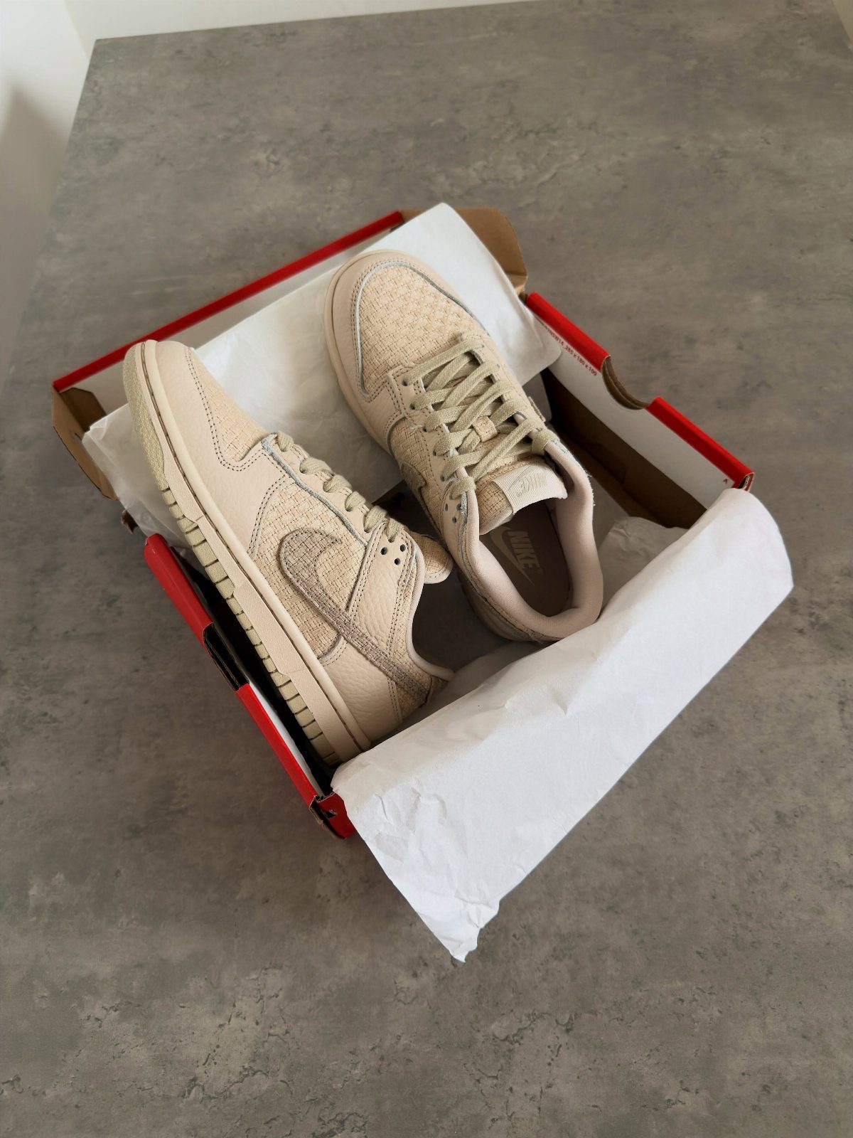 Beige Nike Dunk Low sneakers inside a red and white shoebox on grey stone.