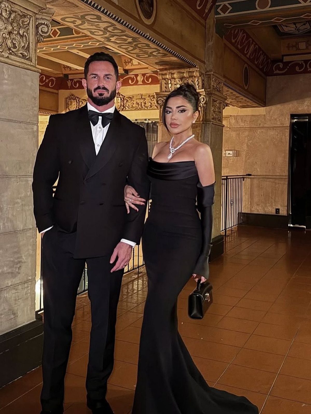 Woman in a long black satin off-the-shoulder gown standing with a man in tuxedo.