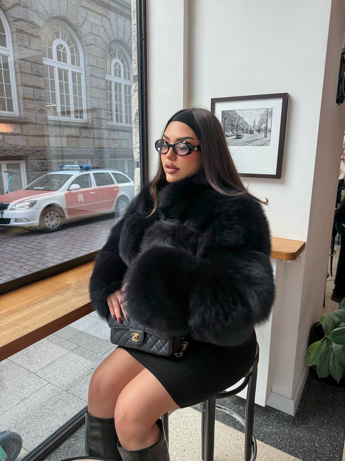 Woman wearing a short black dress and a fluffy black fur jacket sitting indoors.