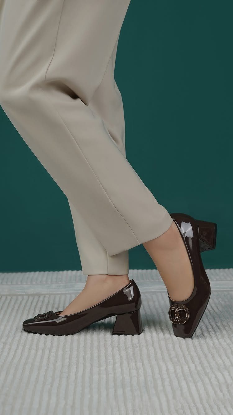 Black patent leather low block heel pumps worn with beige trousers.