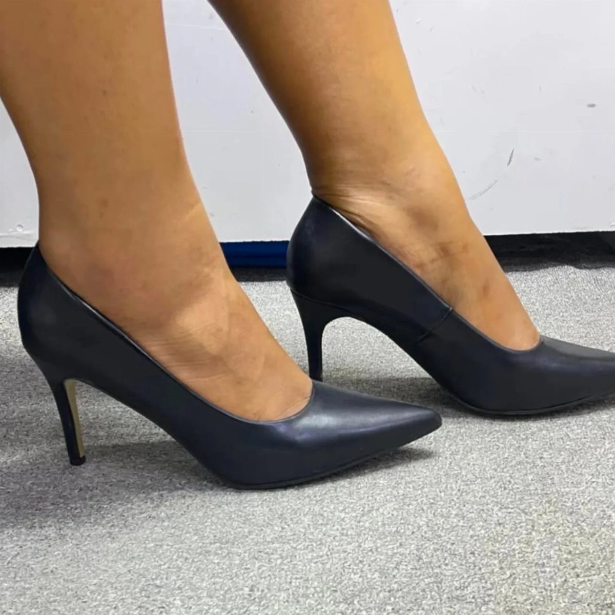 Black pointed-toe pumps with low heels worn on a grey carpeted floor.