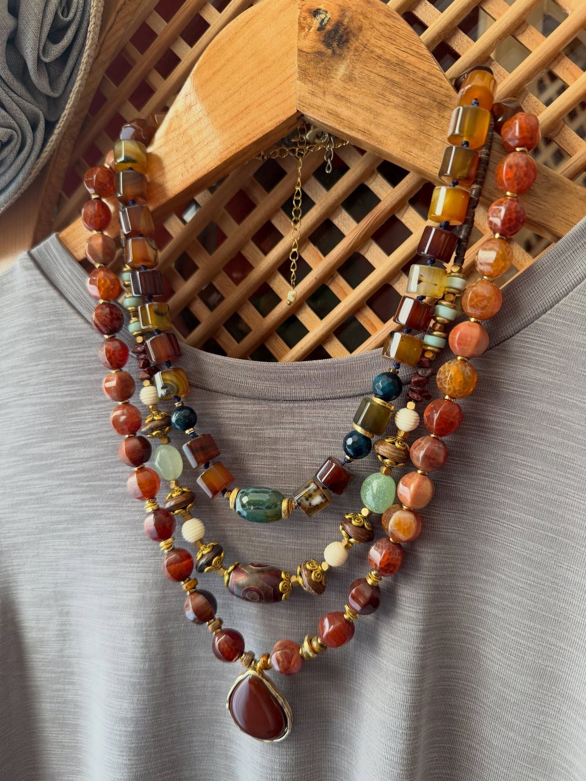 Layered beaded necklaces in earthy tones draped over a grey shirt on a hanger.