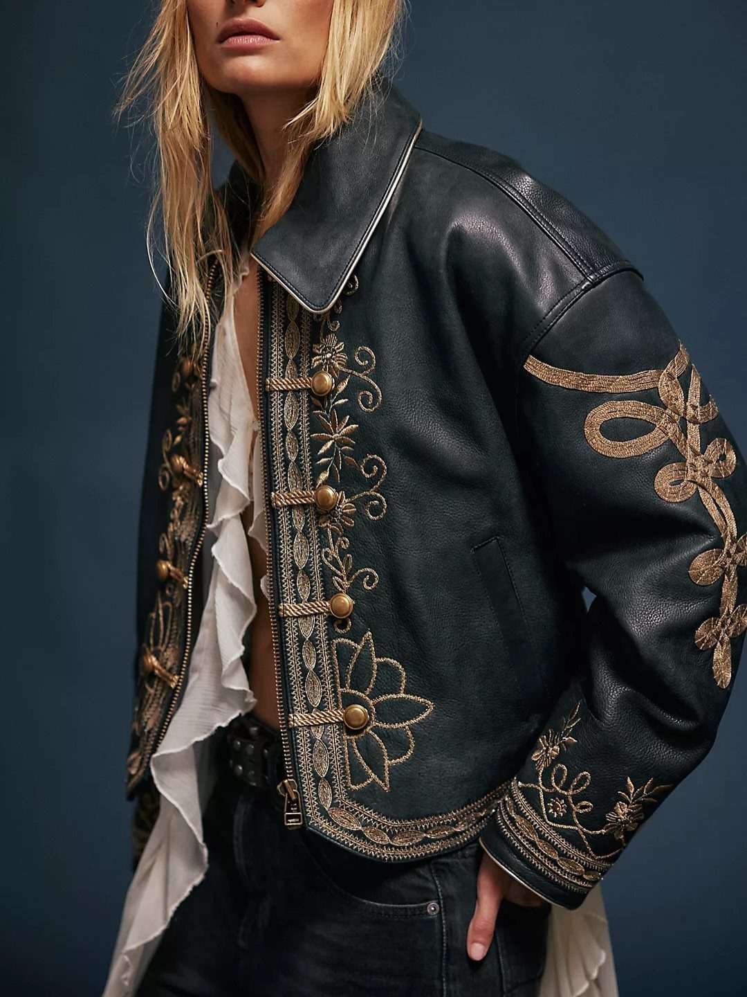 Woman wearing a black leather jacket with gold floral embroidery and white ruffles.