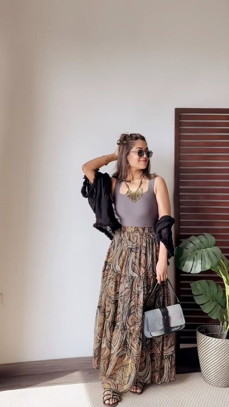 Woman wearing a patterned brown maxi skirt, grey top, and layered necklace indoors.