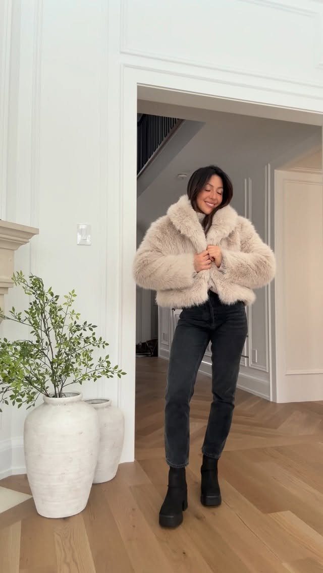 Woman in a fluffy beige jacket and black chunky boots standing in a hallway.