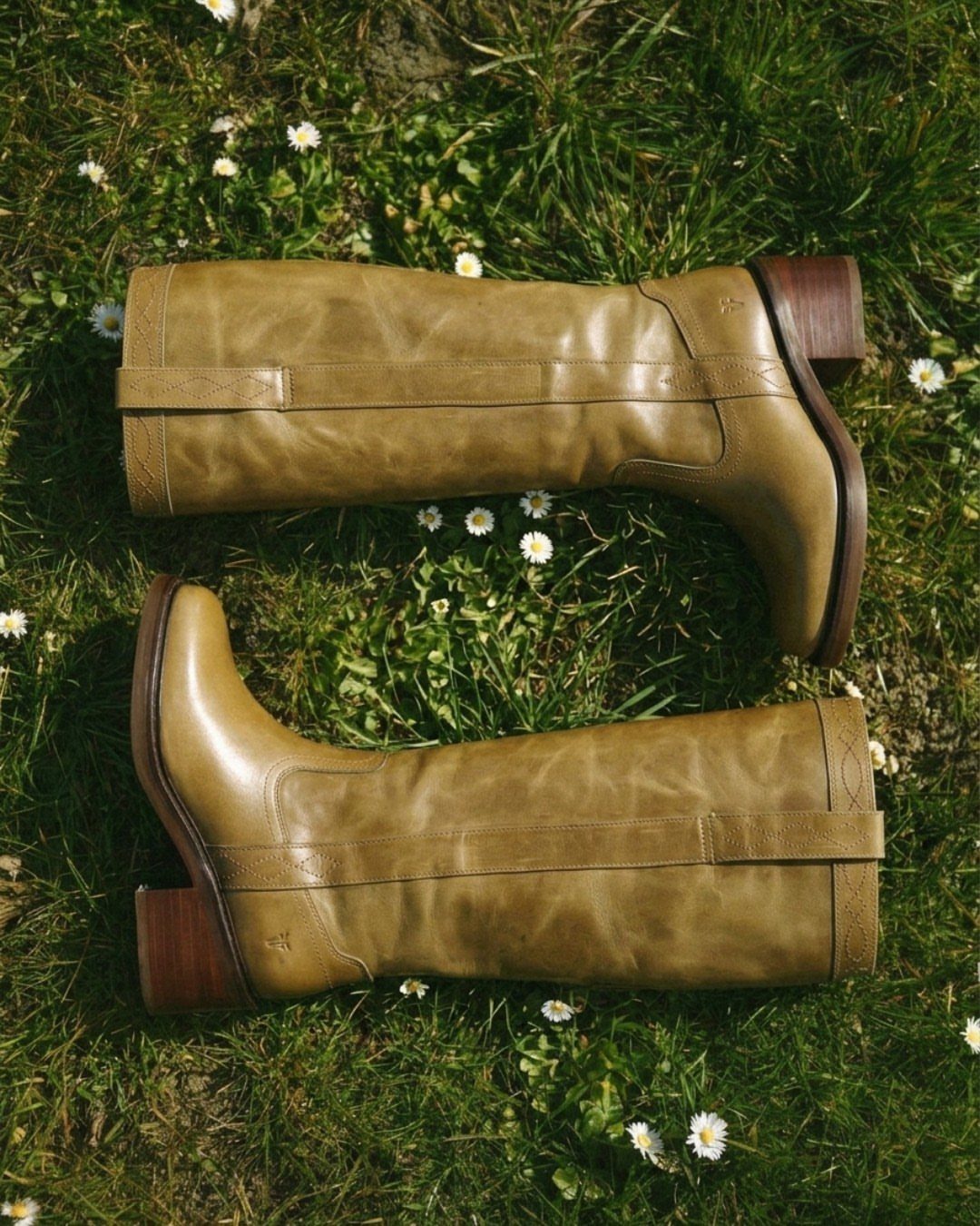 Tan leather mid-calf boots with straps resting on green grass with daisies.