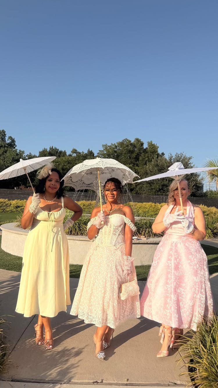 Three women in pastel Regency-style dresses holding white lace parasols outdoors.
