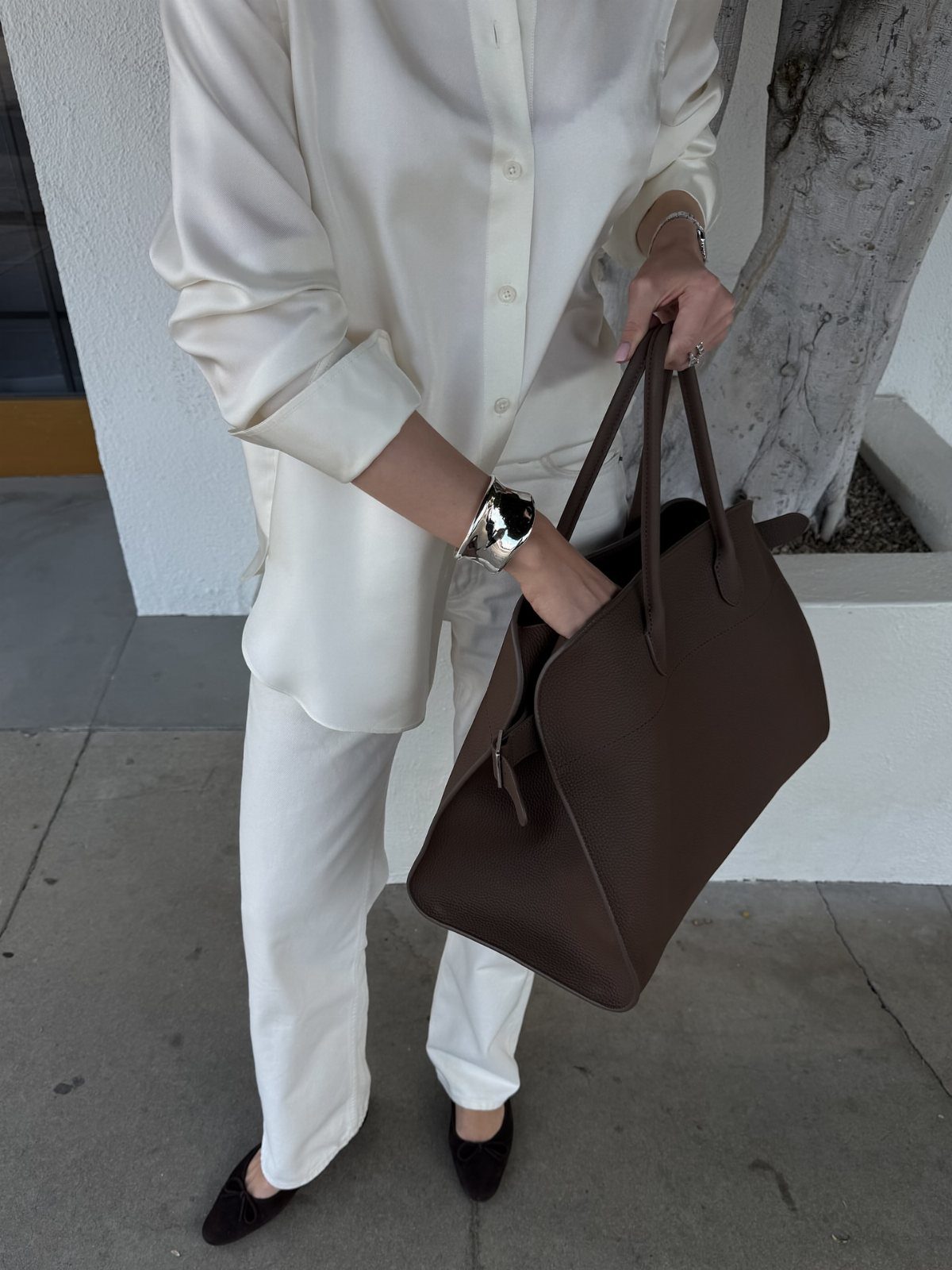 Person in ivory outfit holding a large chocolate brown leather handbag outdoors.