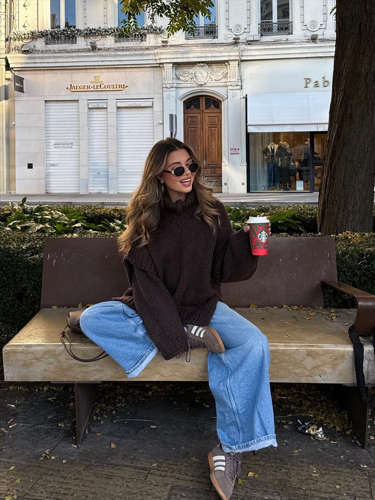Woman in brown oversized sweater and wide-leg blue jeans sitting on a bench.