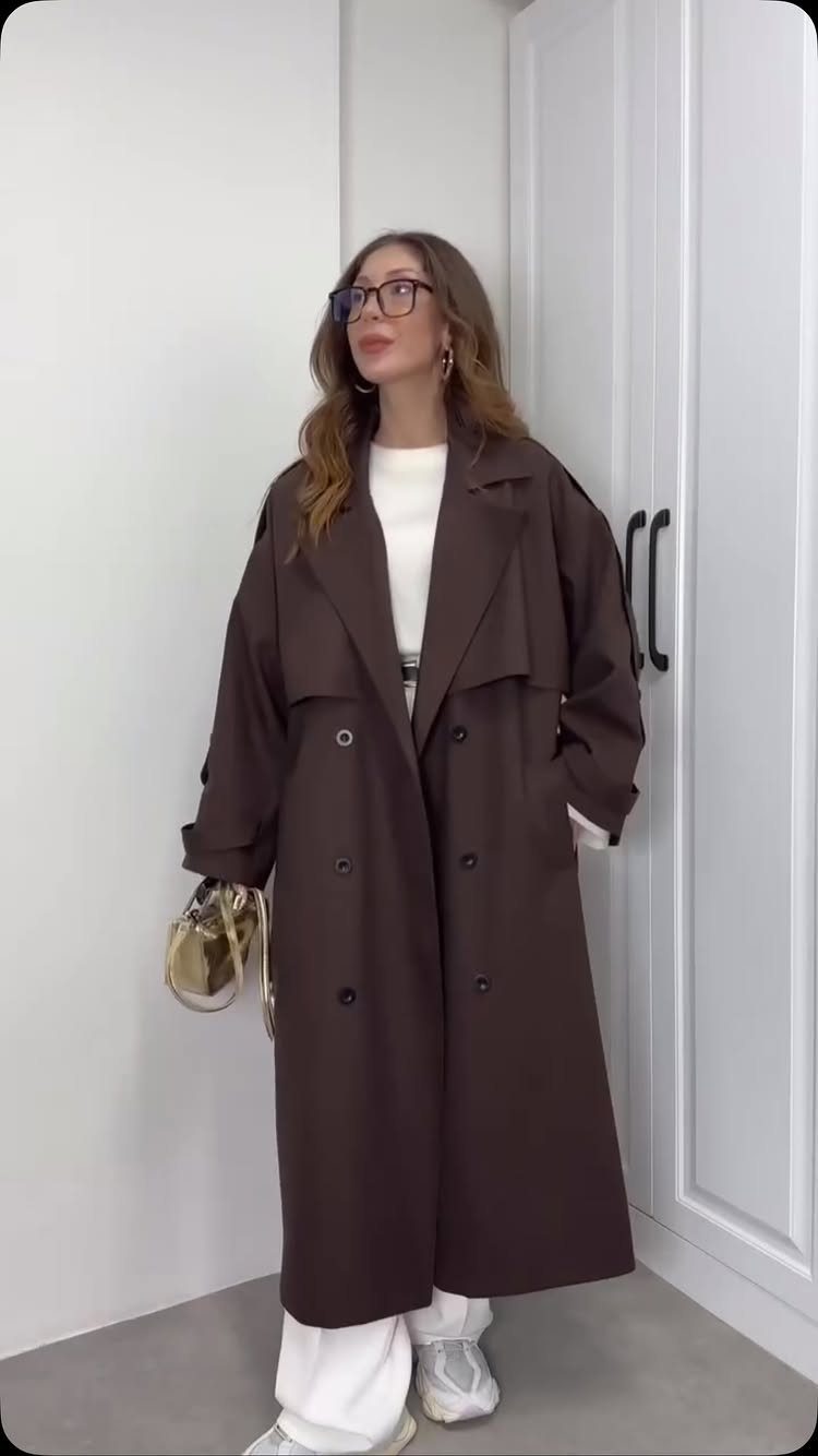Woman wearing a long brown trench coat with white trousers and white sneakers.