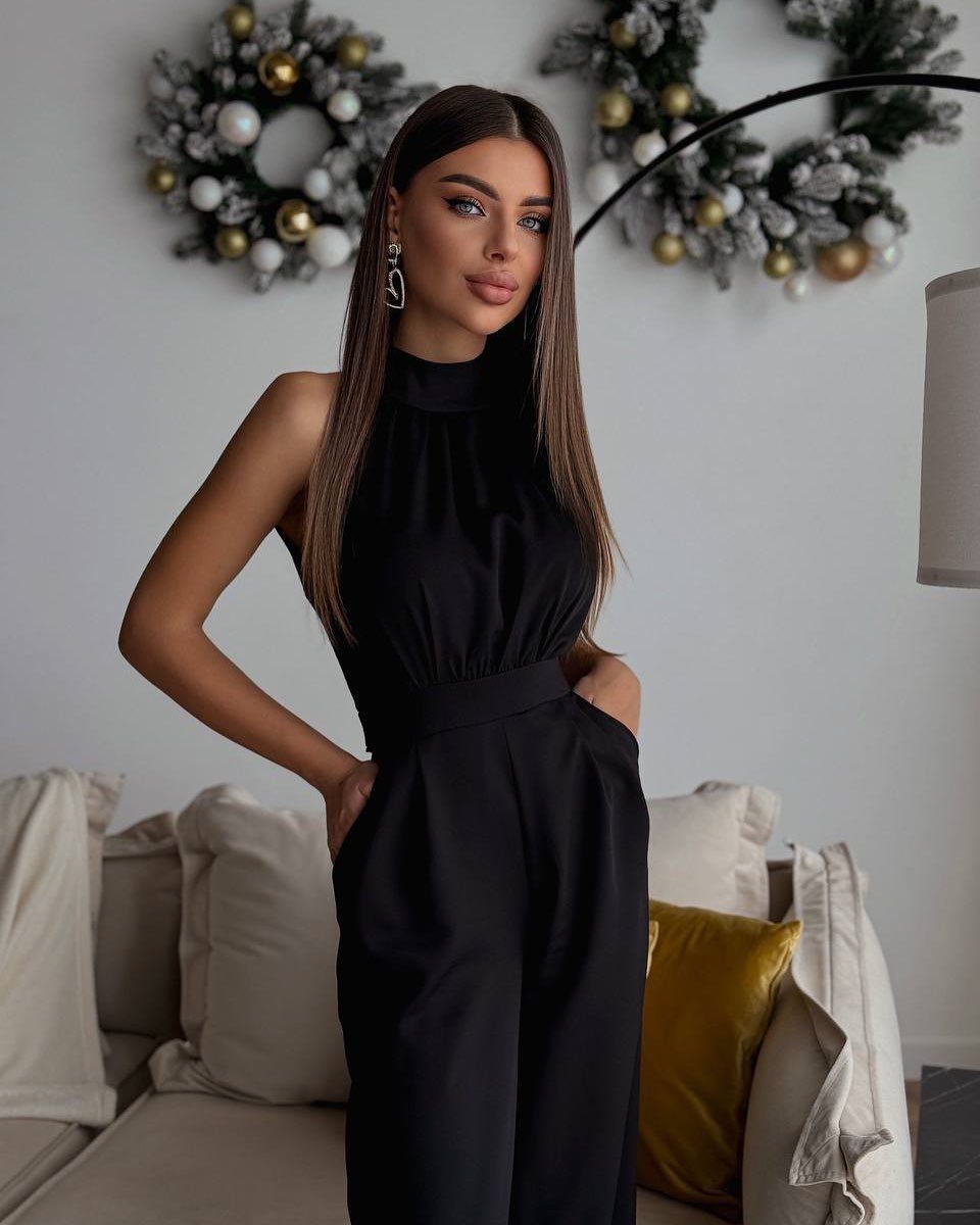 Woman wearing a sleeveless black jumpsuit posing in a room with Christmas wreaths.
