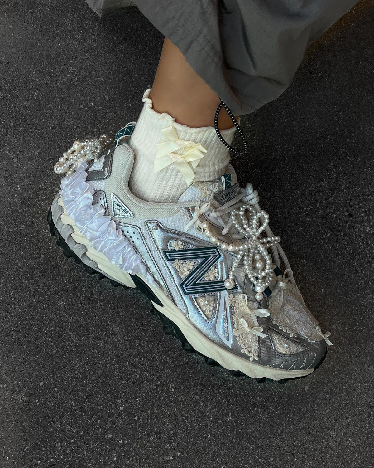 Silver New Balance sneaker with decorative pearl beads and wide white fabric ribbon laces.