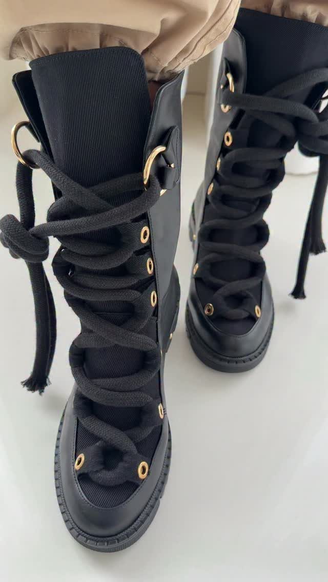 Black high-top canvas sneakers featuring wide black fabric laces and gold eyelets.