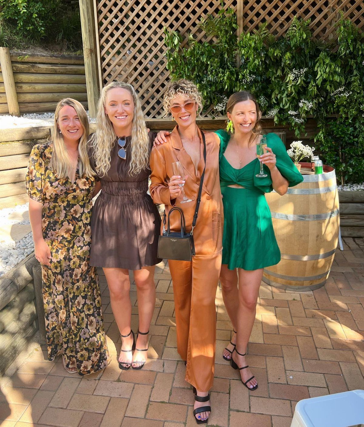 Four women in colorful party dresses and a jumpsuit posing outdoors on a patio.