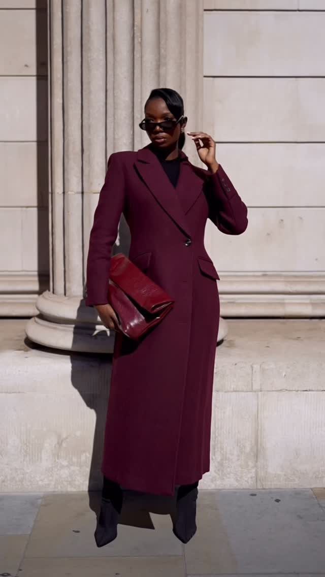 Woman in a long burgundy coat and black boots standing by stone columns.