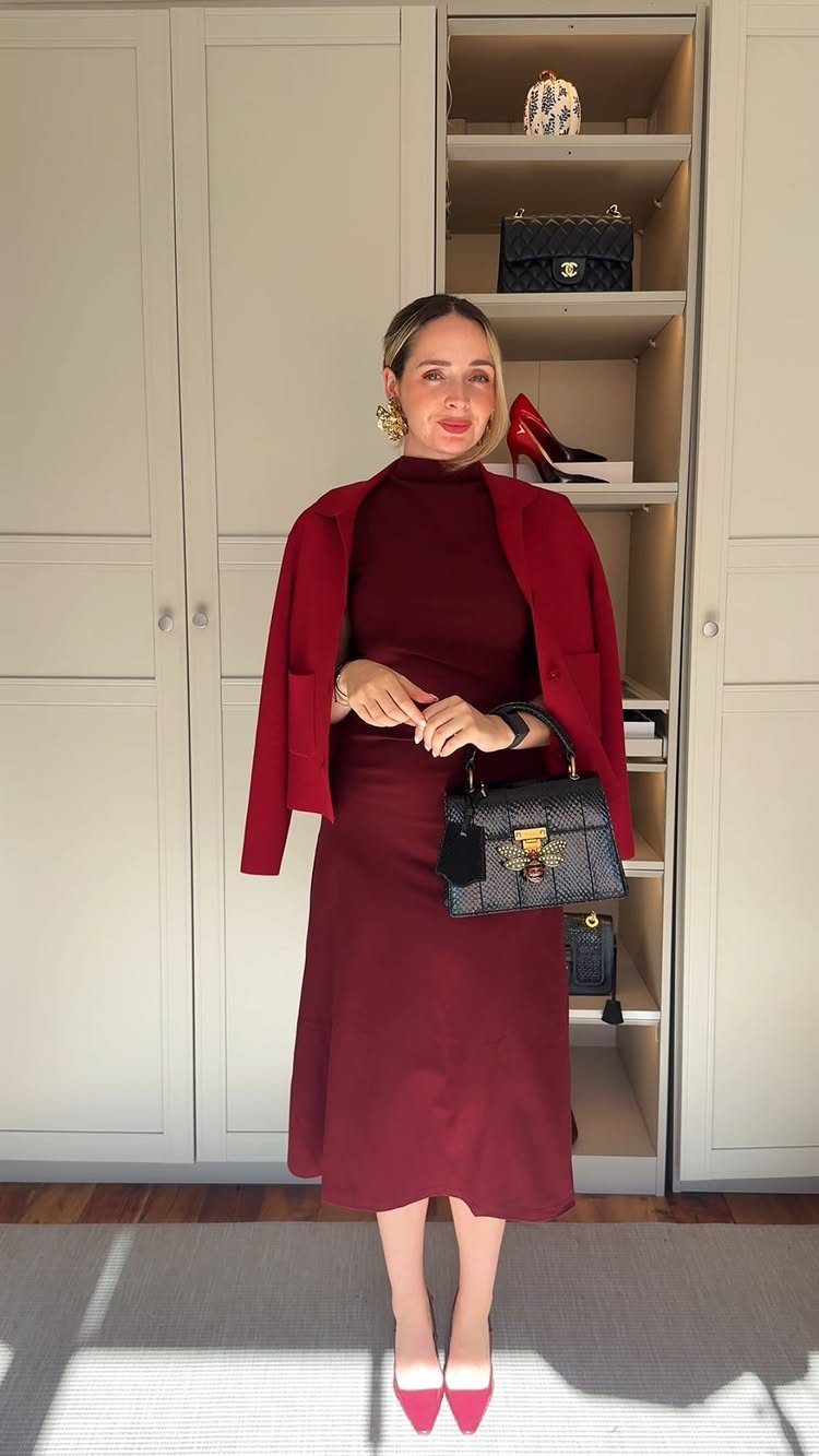 Woman in a monochromatic burgundy dress and blazer holding a black handbag.