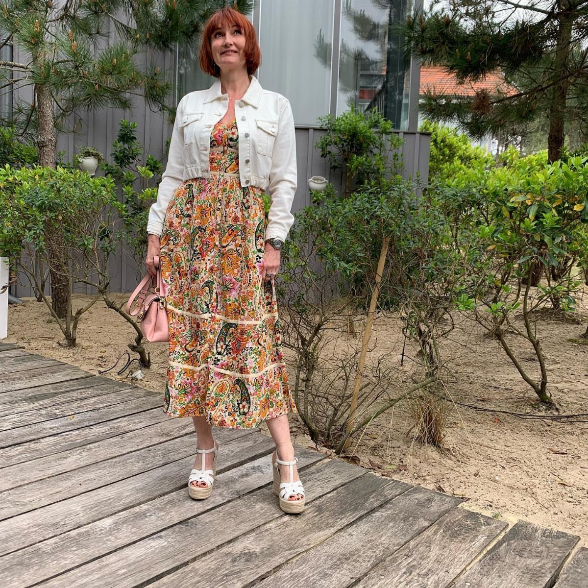 Woman wearing a white denim jacket over a colorful floral midi dress and sandals.