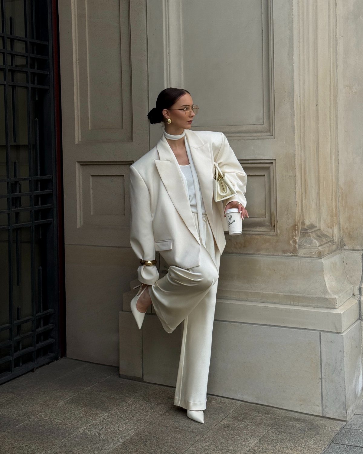 Woman wearing an oversized white blazer and matching white trousers leaning against a wall.
