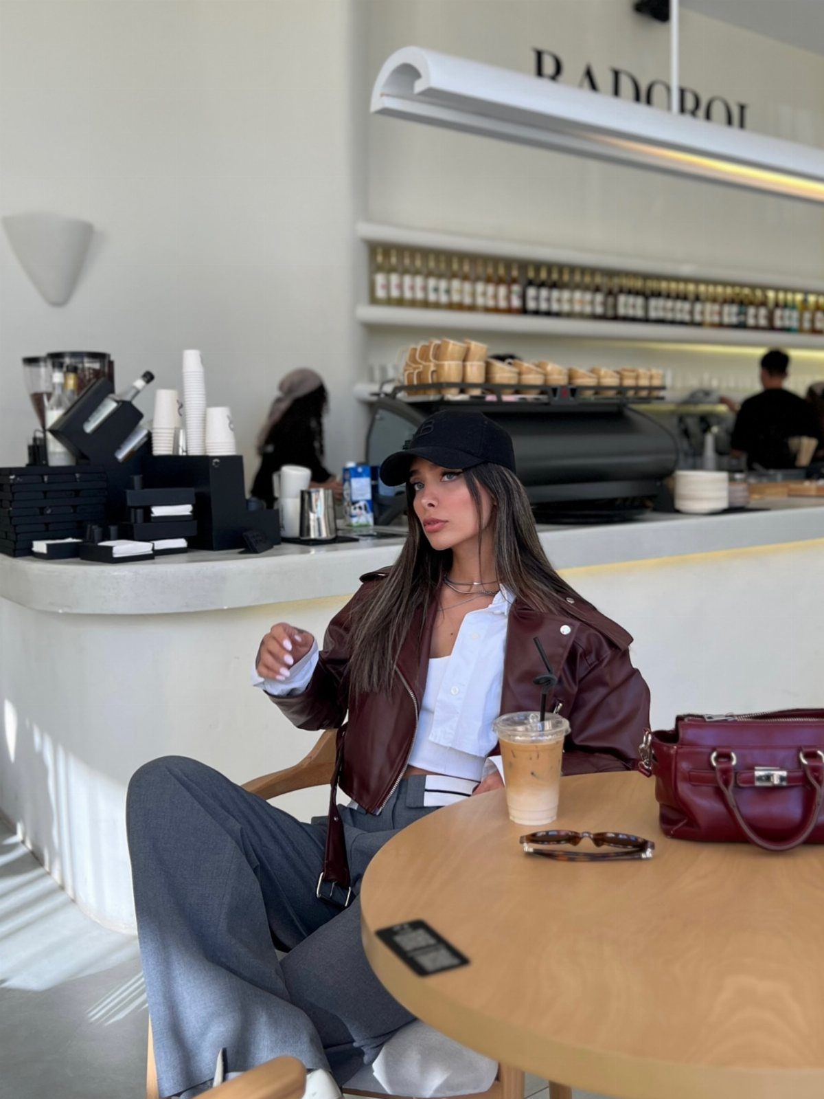 Woman in brown leather jacket, white shirt, and grey trousers at a cafe.