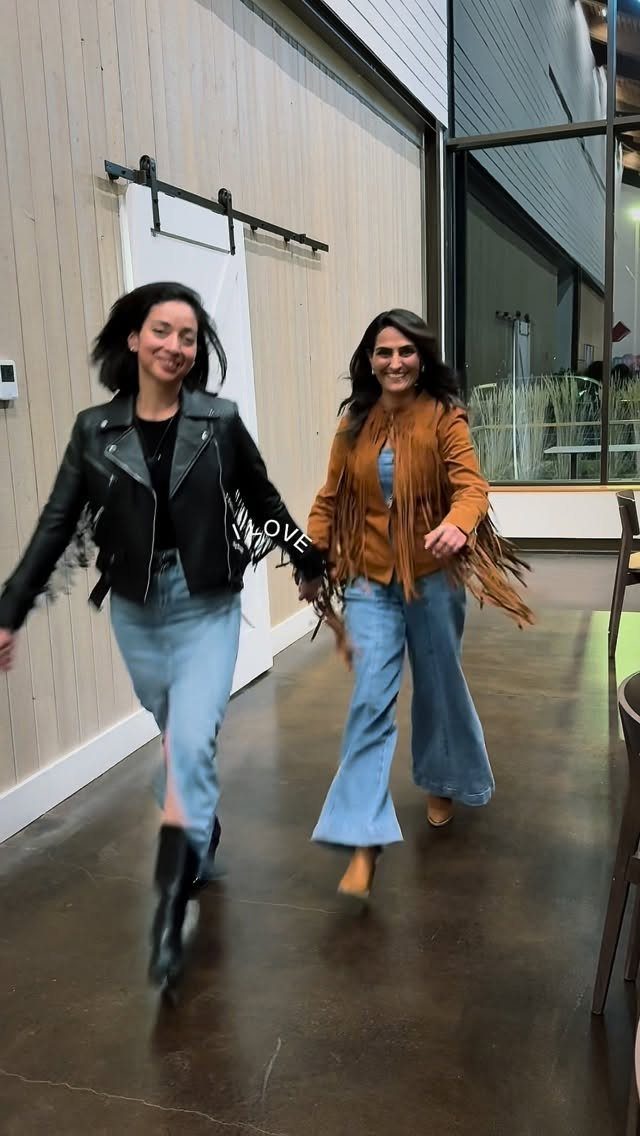 Two women wearing fringed leather jackets and blue jeans walking in a room.
