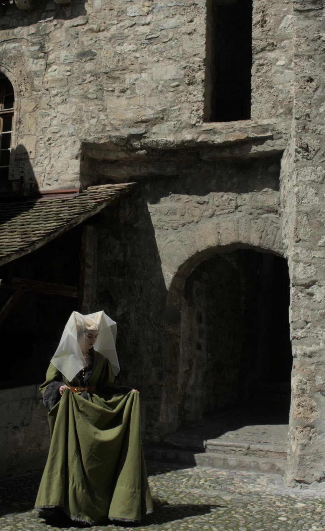 Woman in olive green medieval gown and white hennin veil by stone castle wall