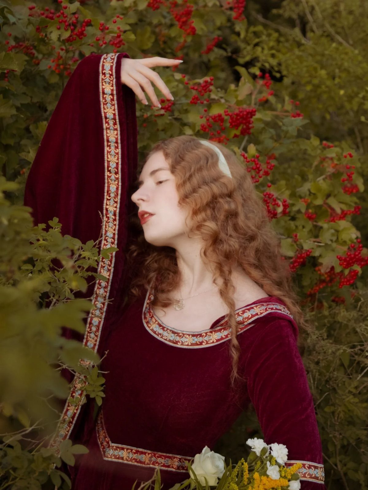 Woman in a red velvet medieval dress with gold embroidery posing in nature.