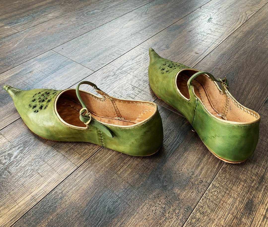 Green leather pointed-toe medieval shoes with decorative perforations on a wooden floor