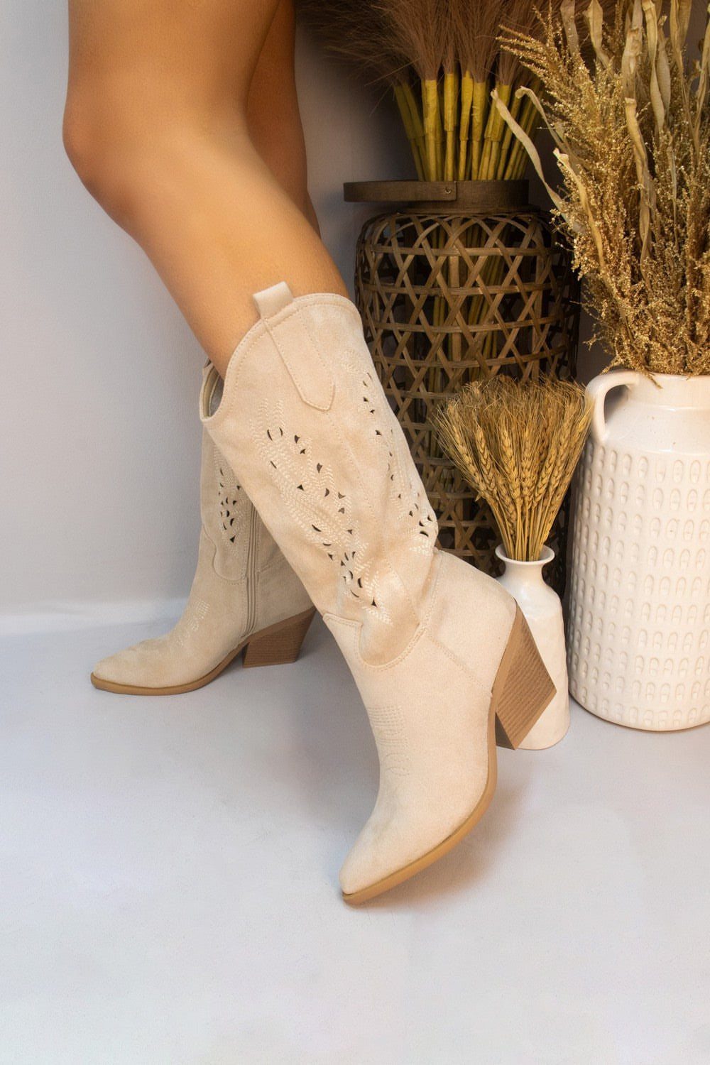 Beige suede mid-calf boots with block heels and decorative cut-outs on a person.