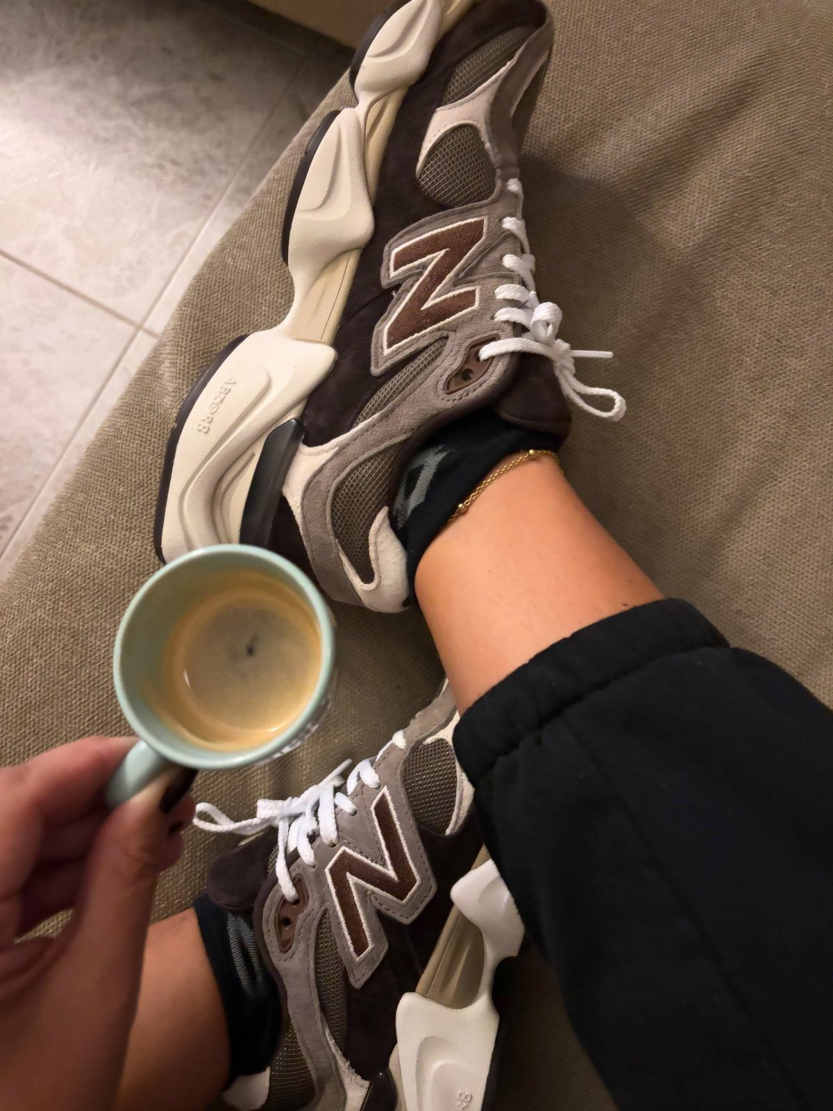 Brown New Balance 9060 sneakers and a cup of coffee on a beige surface.