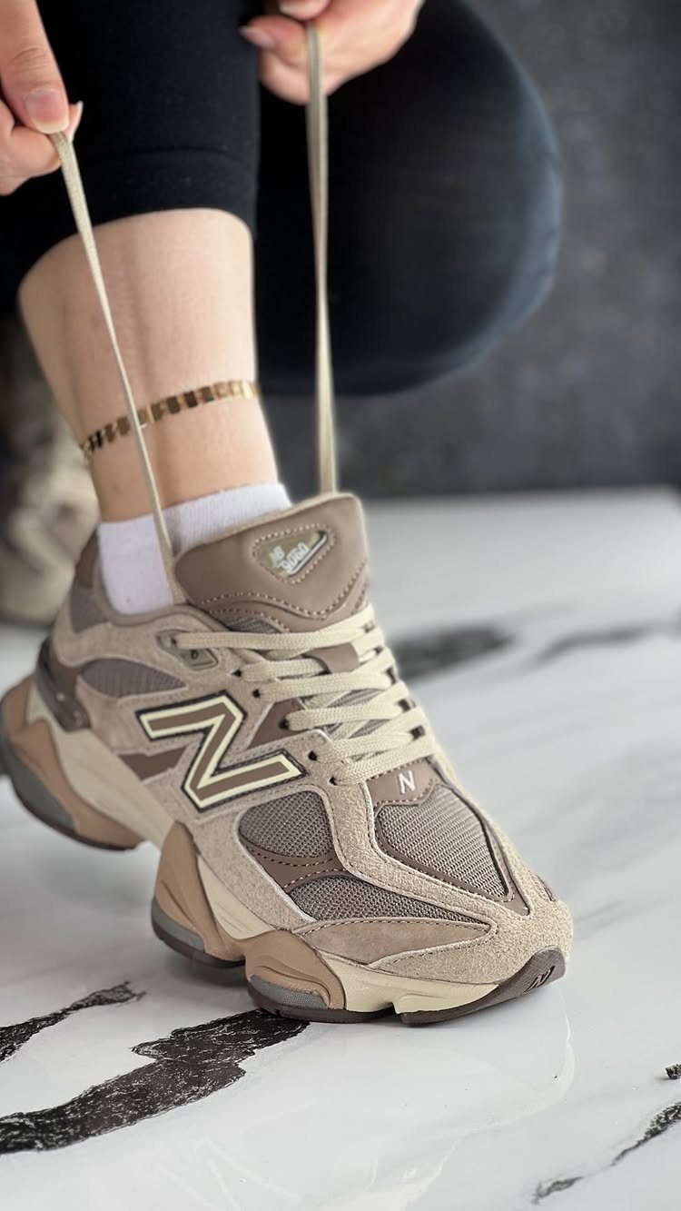 Beige New Balance 9060 sneakers being tied on a white marble floor.