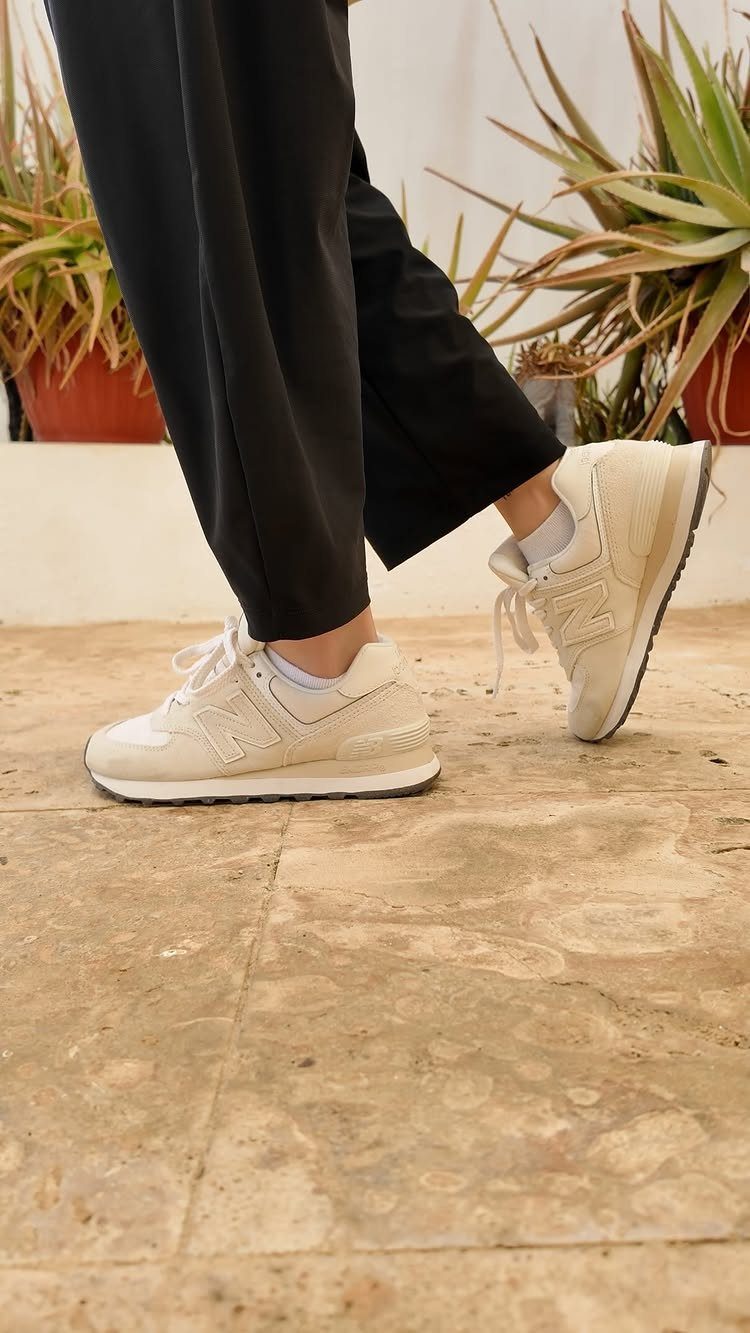 Beige New Balance 574 sneakers worn with black trousers on a stone floor.