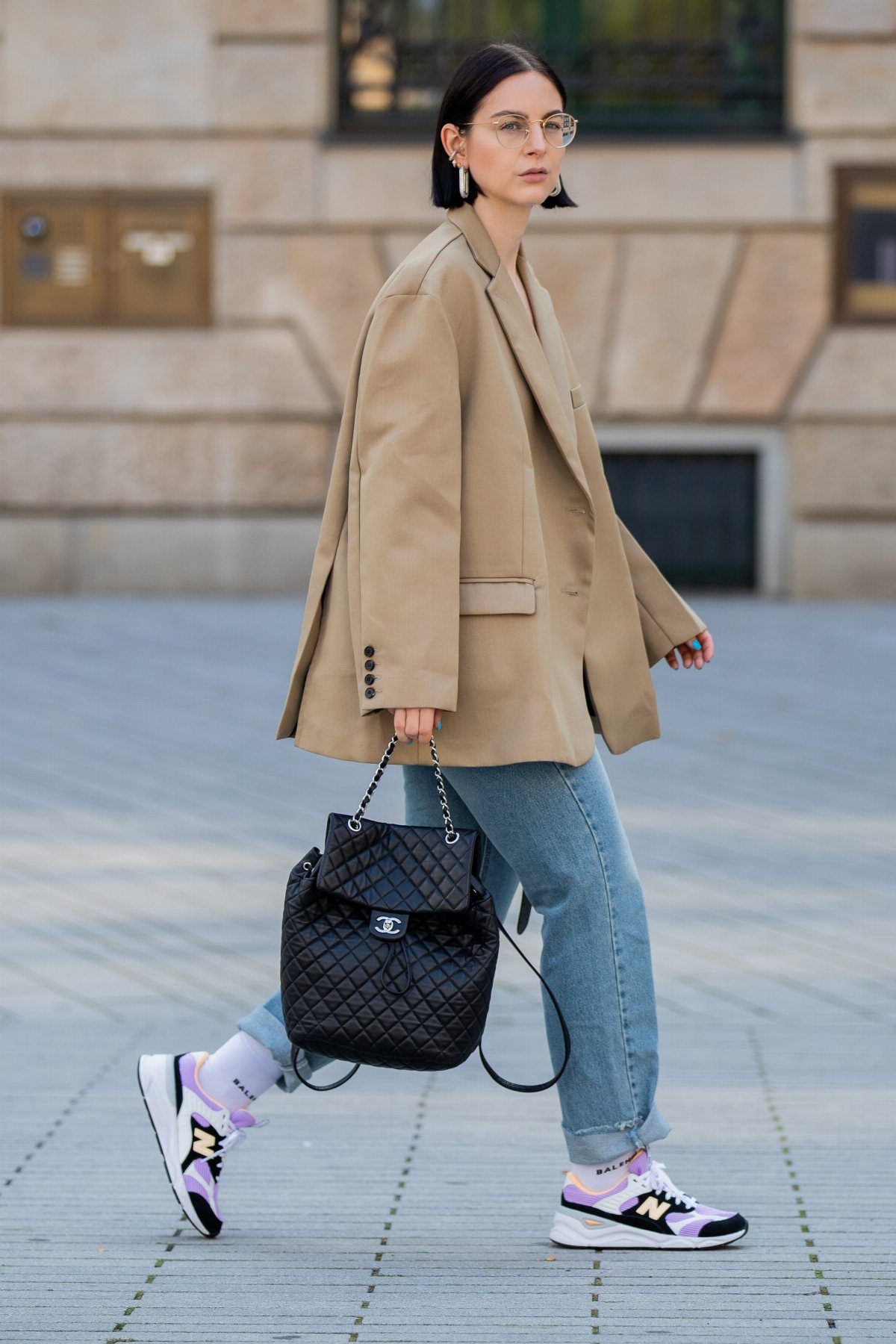 Woman wearing oversized beige blazer, denim jeans, and white New Balance sneakers walking outdoors.