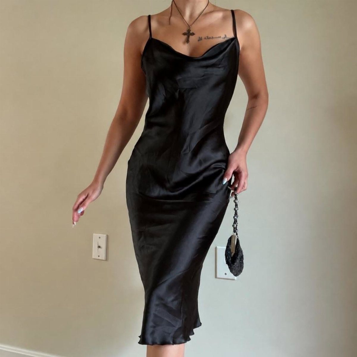 Person wearing a black satin slip dress and cross necklace against a white wall