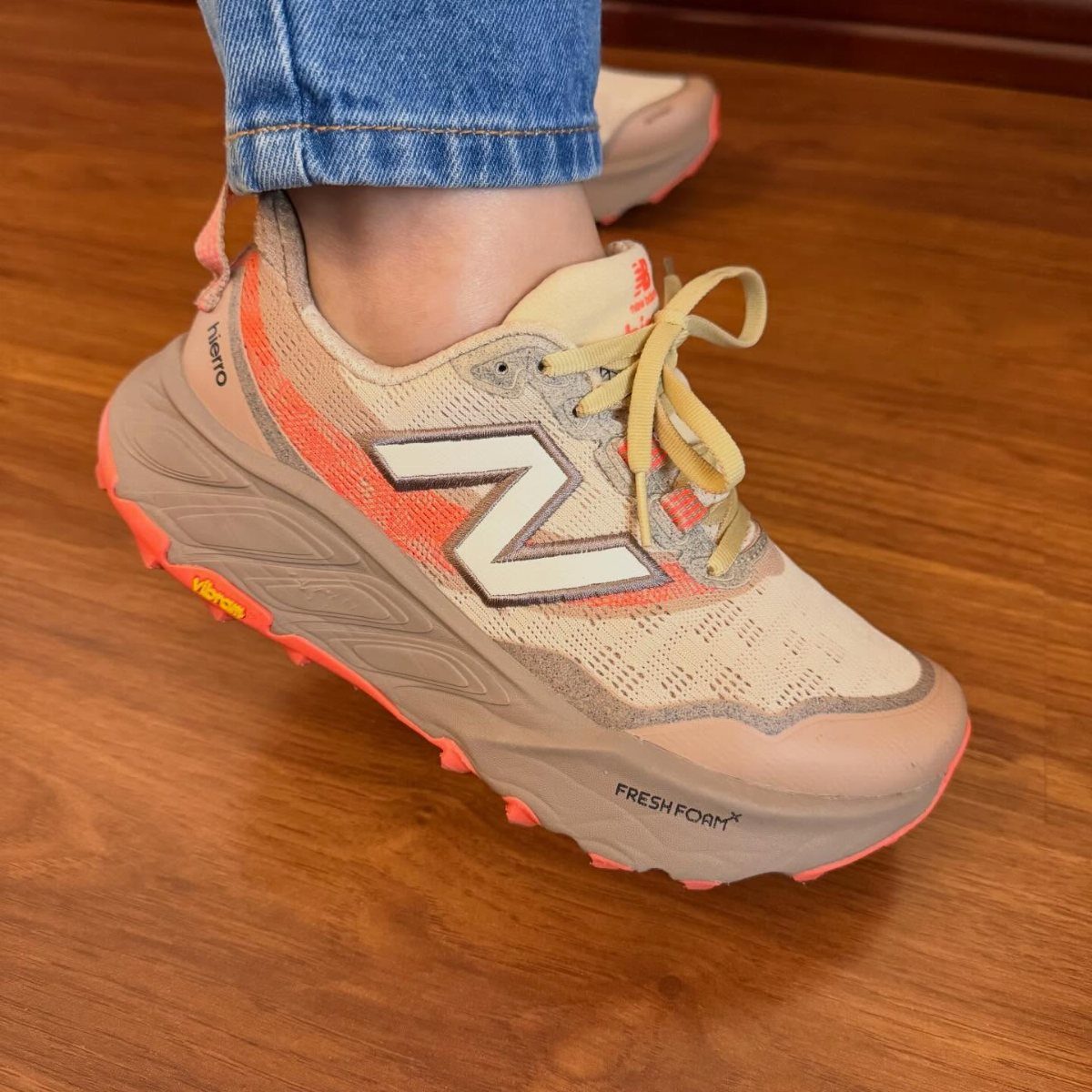 Tan and orange New Balance trail running shoes worn with blue jeans on wood.