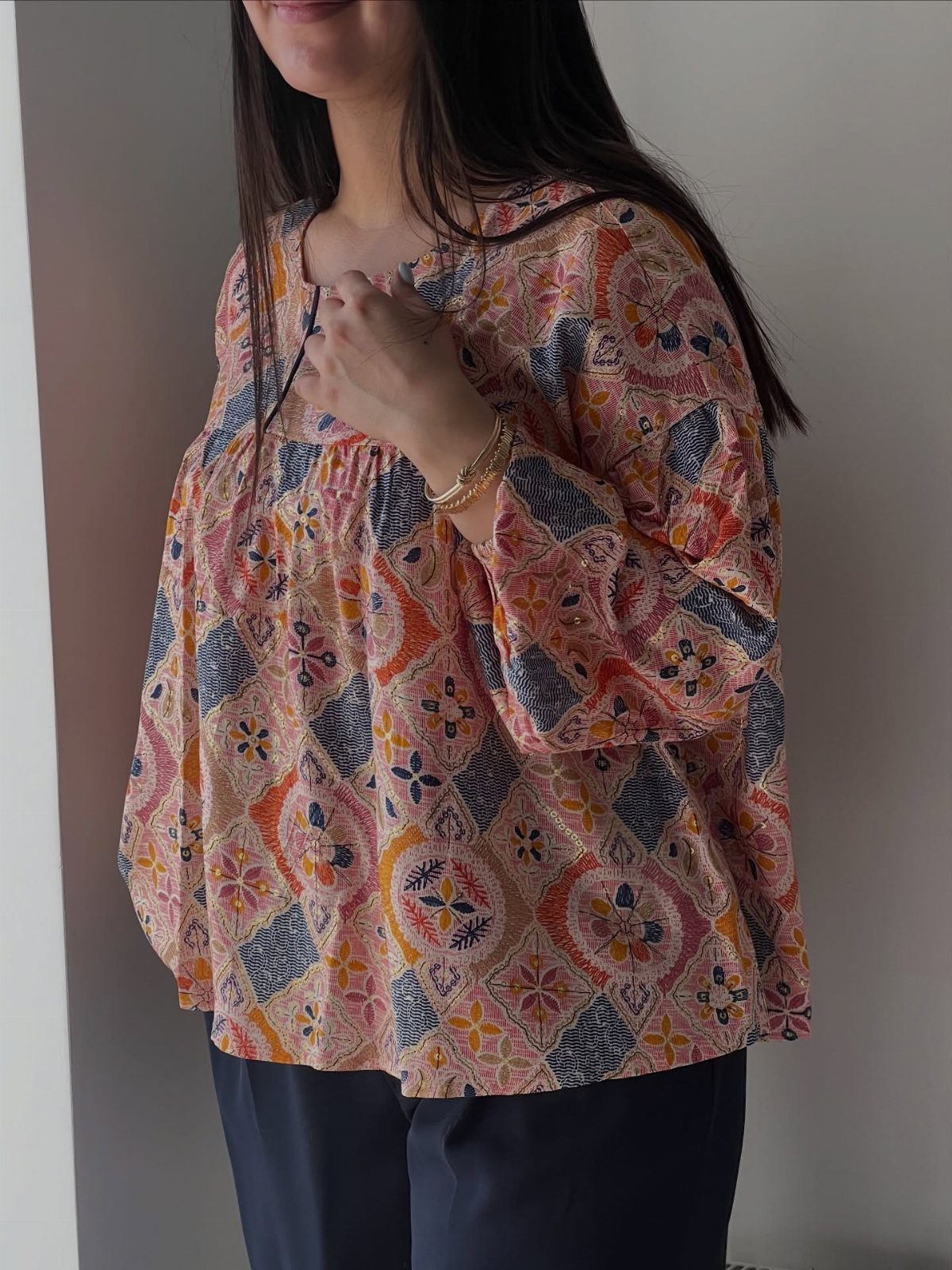 Woman wearing a billowy patterned orange and blue blouse with navy trousers.