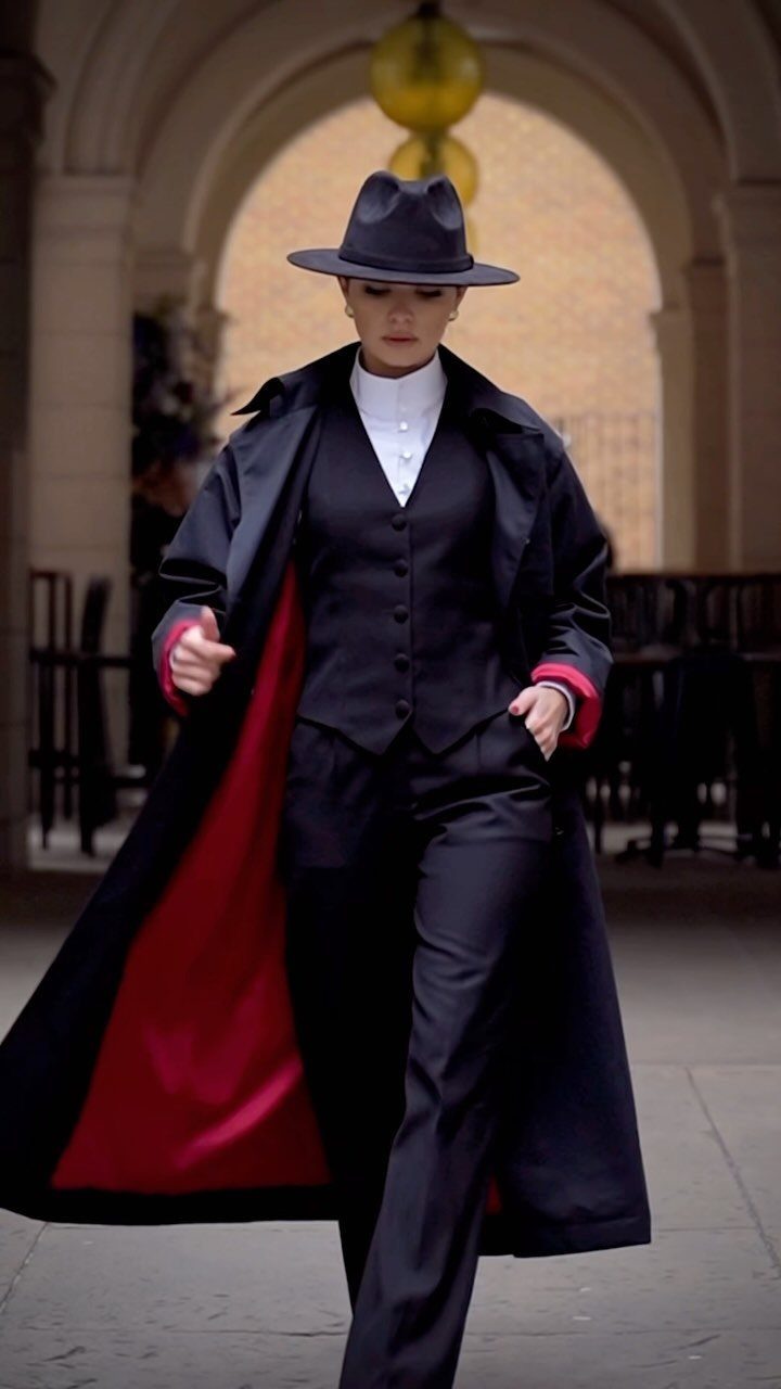 Person in a black tailored suit and fedora walking through a stone archway.