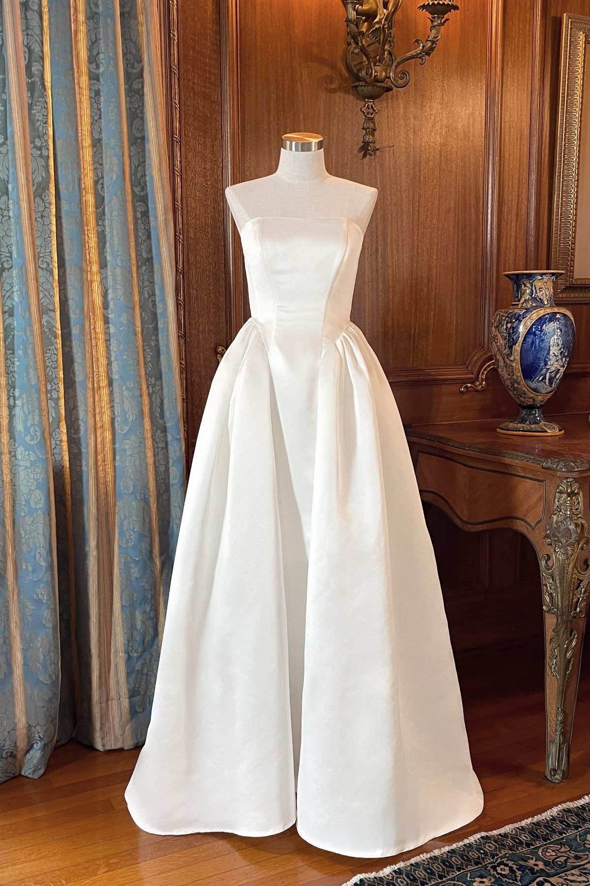 White satin strapless gown on a mannequin in a vintage wood-paneled room.