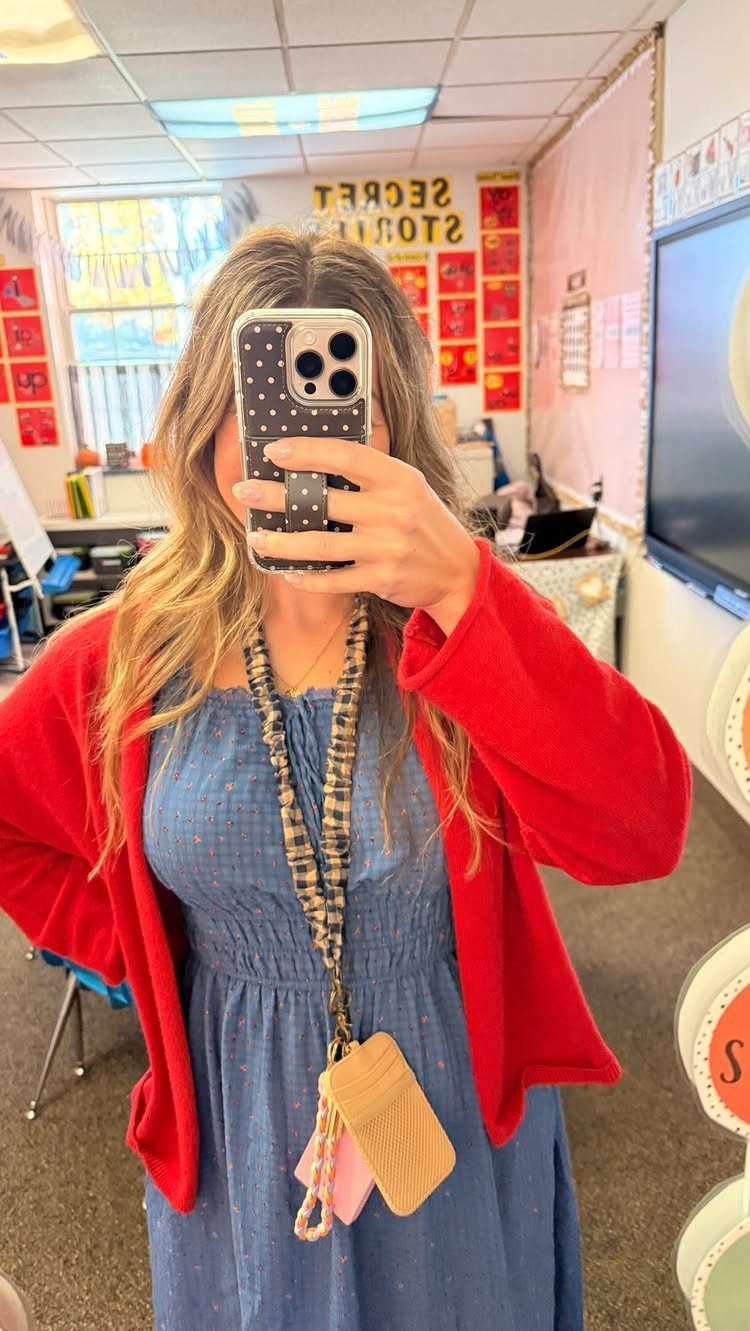 Woman in a red cardigan and blue dress taking a mirror selfie in classroom