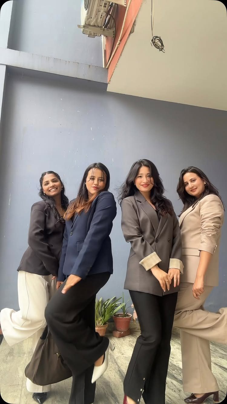 Four women in coordinating neutral-toned business blazers and trousers posing against a blue wall.