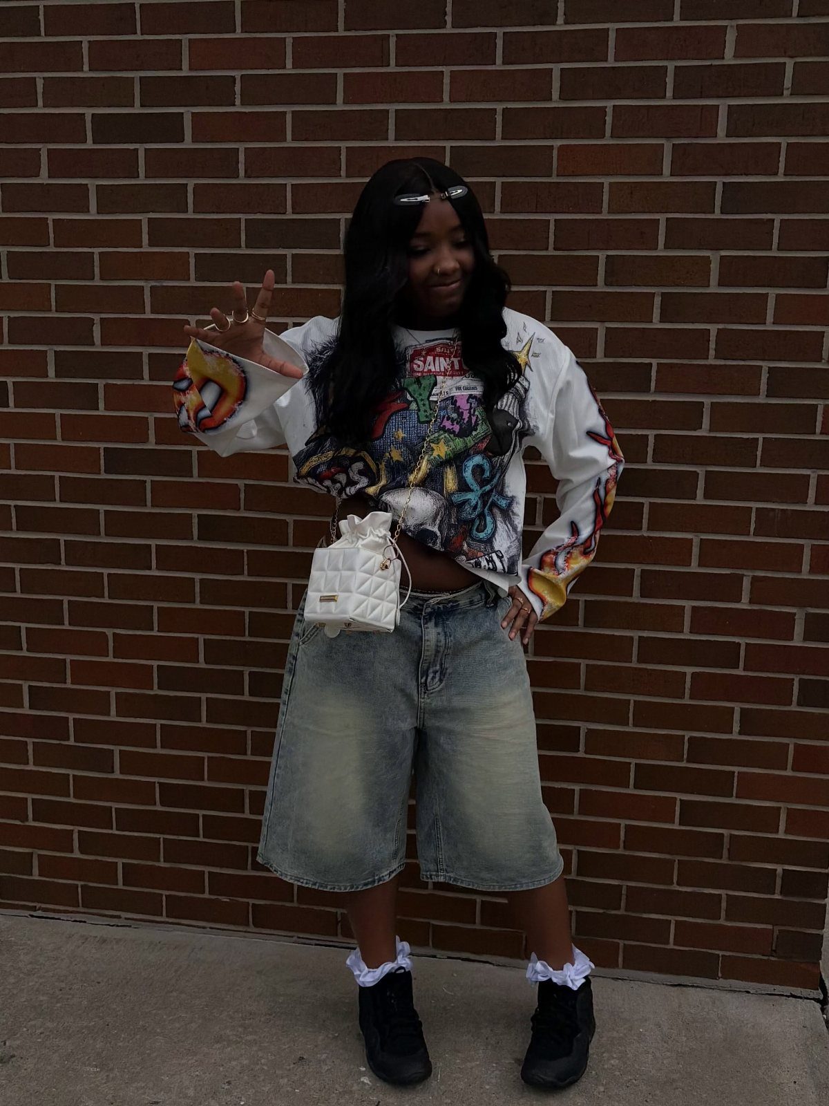 Young woman posing in a graphic shirt and denim shorts against brick wall