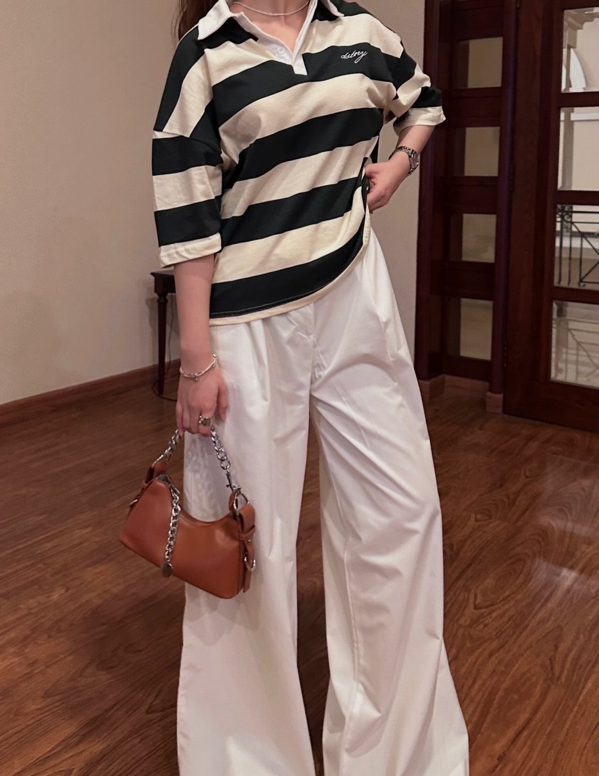 Person wearing a striped polo shirt and wide-leg white trousers holding a brown handbag.