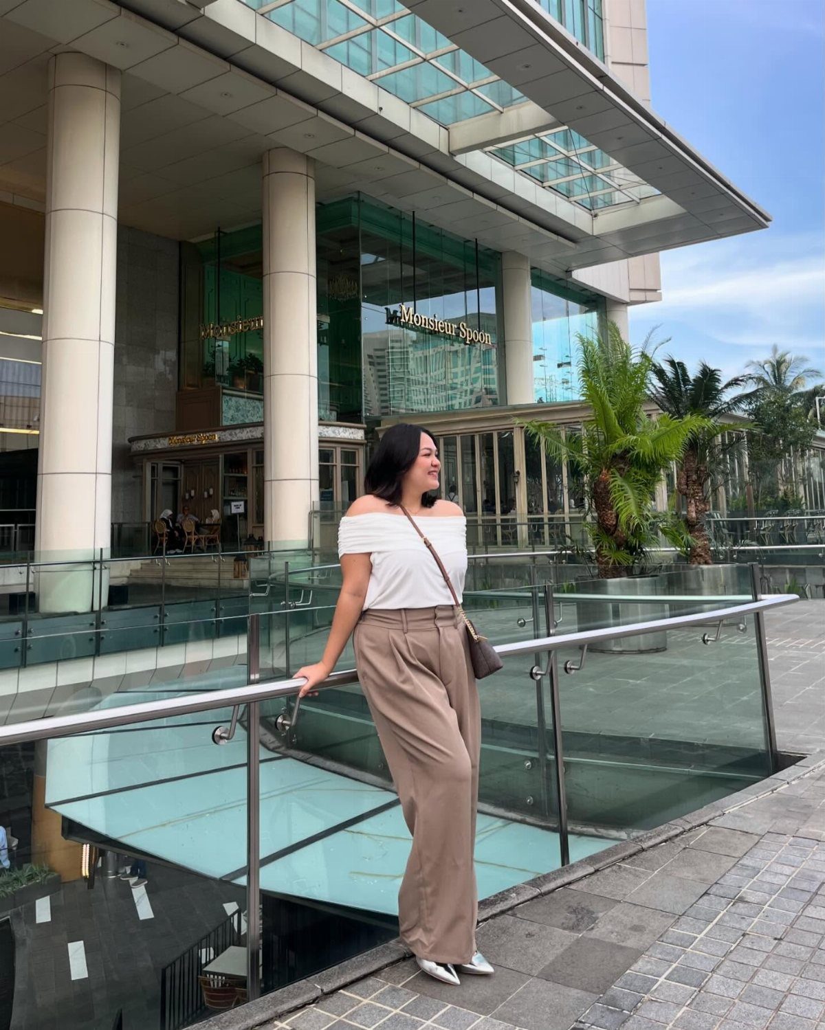 13 Plus Size Brunch Outfit Ideas for Every Style 7 Woman in white off-the-shoulder top and tan wide-leg trousers standing by glass railing